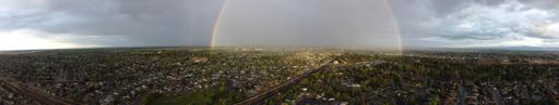A wide pantographic image of a town in California's Central Valley. The skies are cloudy and there is a rainbow with light at the center.