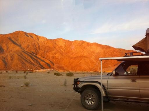 In the foreground is  Toyota Land Cruiser 80 series with awning and RTT deployed. Behind it the desert stretches into the background, to a low train bridge, and then there is a small mountain ridge that is bathed in orange light alpenglow from the sunrise.