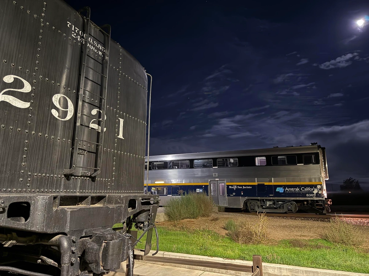 A tale of two trains. In the foreground is the back of the Modesto Bee steam locomotive with the number 2921 painted in white. In the background is the front of a modern Amtrak trail. Cloudy skies are illuminated in the darkness behind.