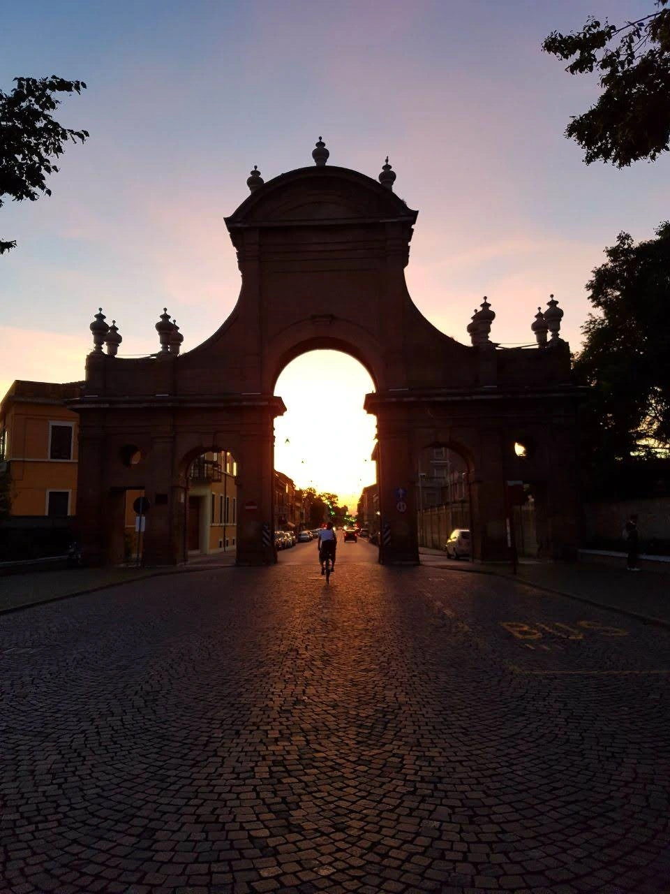 An image of the Prospettiva della Giovecca in Ferrara, Italy during sunset. The large archway frames the setting sun, casting a warm orange glow on the cobblestone street. A silhouette of a person on a bicycle is seen approaching the arch, with buildings lining the sides of the street. The sky is a gradient of purples and pinks, complementing the historic architecture.