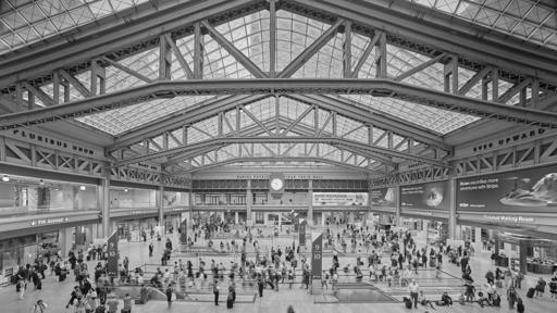 A modern urban passenger rail station hall. Passengers mill about around escalators that descend to unseen tracks below. Exposed steel beams support a skylight roof.