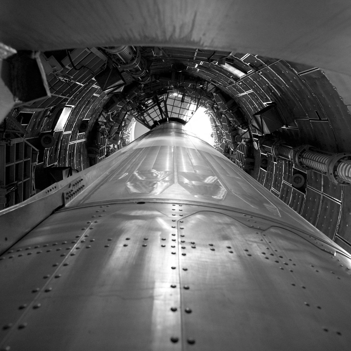 Looking up from the bottom of a missile pointed up inside a cramped underground silo, which is lined with retractable platforms and various apparatus.