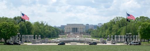 A photo of Lincoln memorial seen from the Washington monument over the reflecting pond. A couple of US flags fly on each side, small black MIA flags beneath them. The memorial is on the center over the pond, the sky is cloudy and blue, dense trees flank the sides of the pond.
