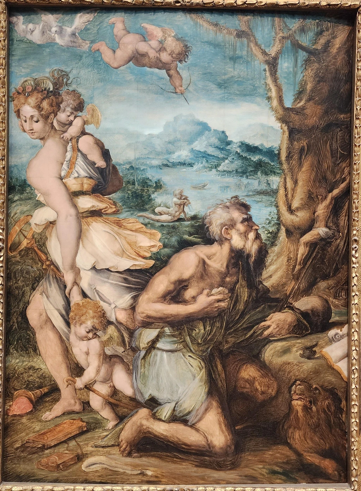 Jerome dressed in rags and kneeling looking at a crucifix. A lion is lying under his knee. Behind him Venus is seen walking away, dragging Cupid along. in the background is a body of water in front of a mountain.