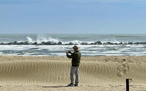 A lone figure - me - stands on wind-swept sand, capturing the restless sea with a phone. Waves break against ancient rocks, their white spray frozen mid-dance beneath a calm blue sky.