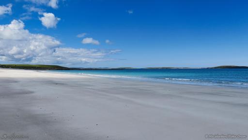 A colour photo of a beach scene. Wide sands stretch out before the viewpoint and, on the horizon, a line of low green islands. The turquoise sea is ruffled by small waves which break gently onto the beach but otherwise it's quite still and clean. The sky is clear apart from a bank of cloud over the land on the left, and a patch of sunlight is brightening the shell sand along the strand.