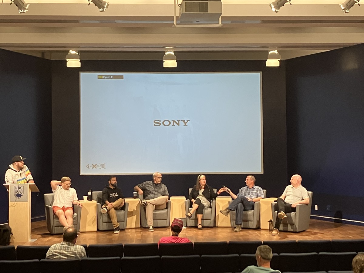 A panel discussion is taking place in a lecture hall at UBC. Six people are seated on stage in armchairs, with one person standing at a podium on the left. The screen behind them displays the word “SONY” in the center. The audience is seated in rows facing the stage.
