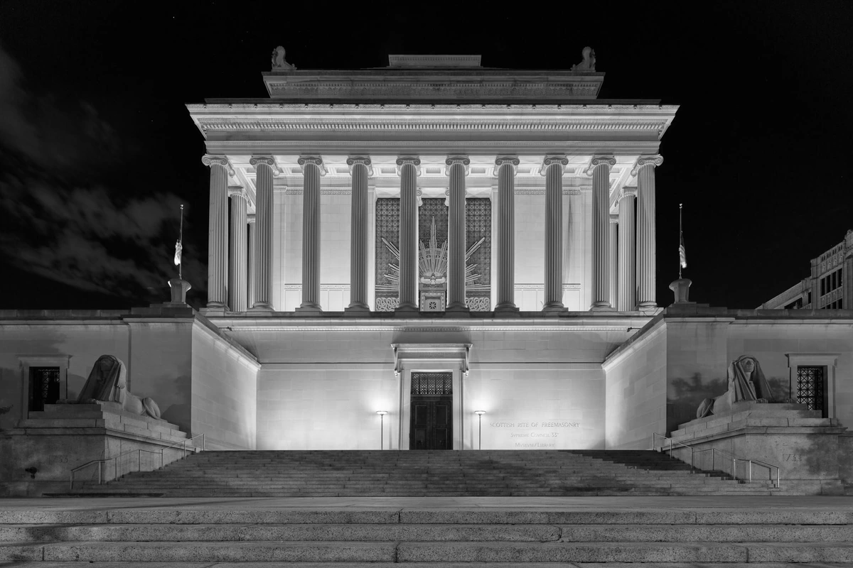 A large Masonic temple at night, with Ionic columns, ornate features, flanked by human lions statuary.