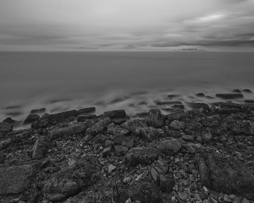 A rocky shoreline with gentle waves lapping at it. In the distance at right is a small island, somewhat enveloped by fog, with a prominent water tower and industrial-type buildings.