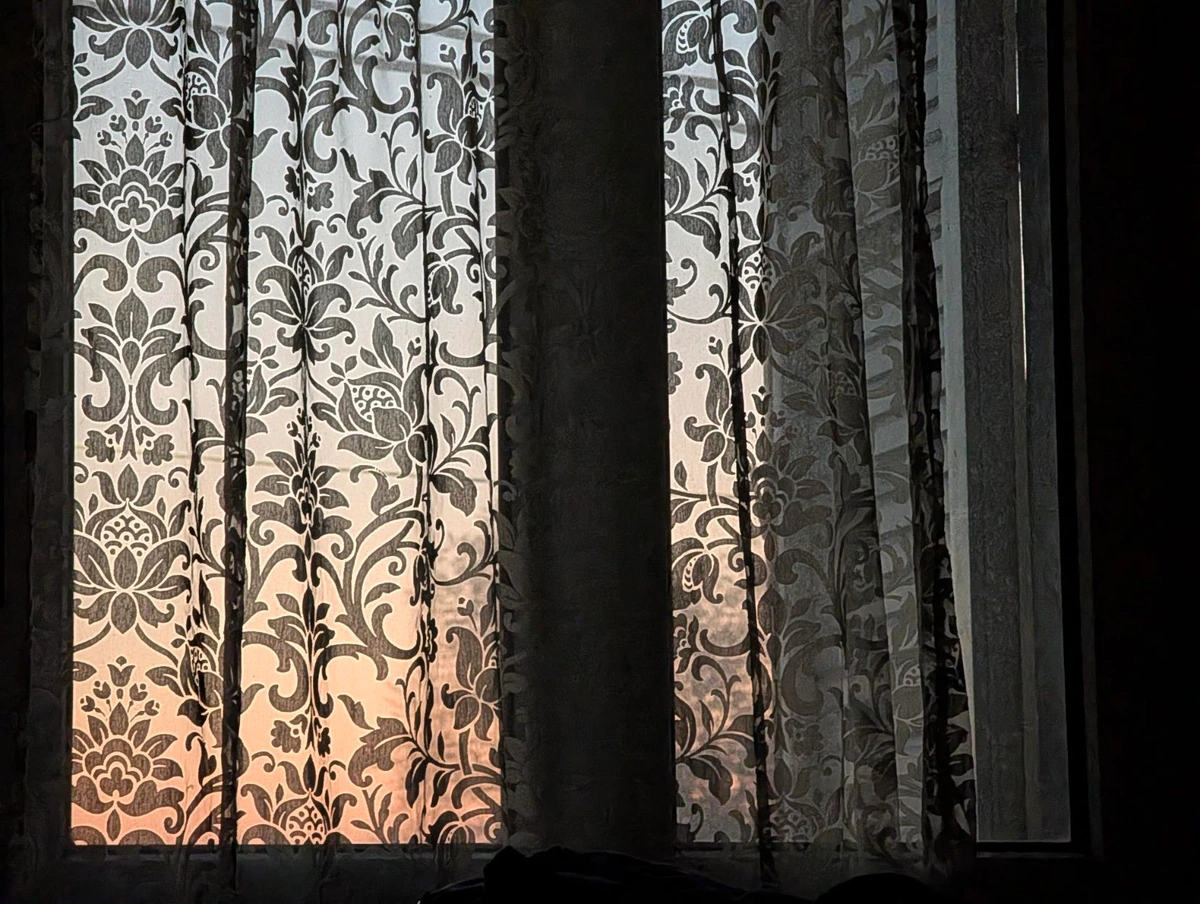 Semi transparent patterned curtains hang in front of a window before sunrise, with soft early light glowing through the fabric and revealing floral motifs while the room remains mostly dark.