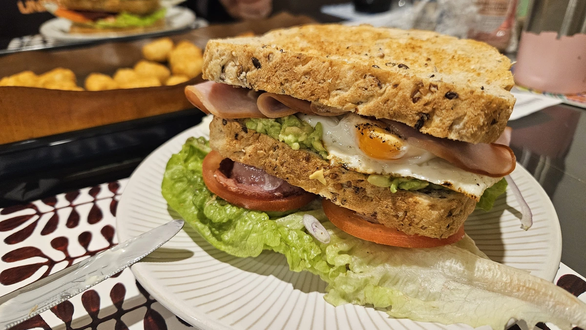 Triple decker sandwich with ham, fried egg, smashed avocado, bacon, tomatoes, red onion, lettuce, mayo, mustard. All on a white plate. Baking tray of tater tots in the background. 
