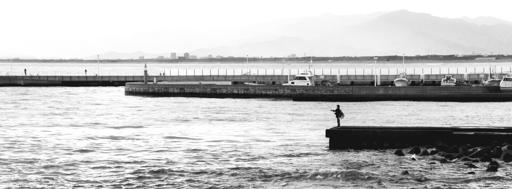 The image shows a black and white scene of a waterfront with a calm body of water, possibly a sea or lake. There is a person standing on a pier or dock extending into the water, possibly fishing. The background includes a barrier, some boats docked along the edge, and distant mountains. The atmosphere is tranquil and serene.