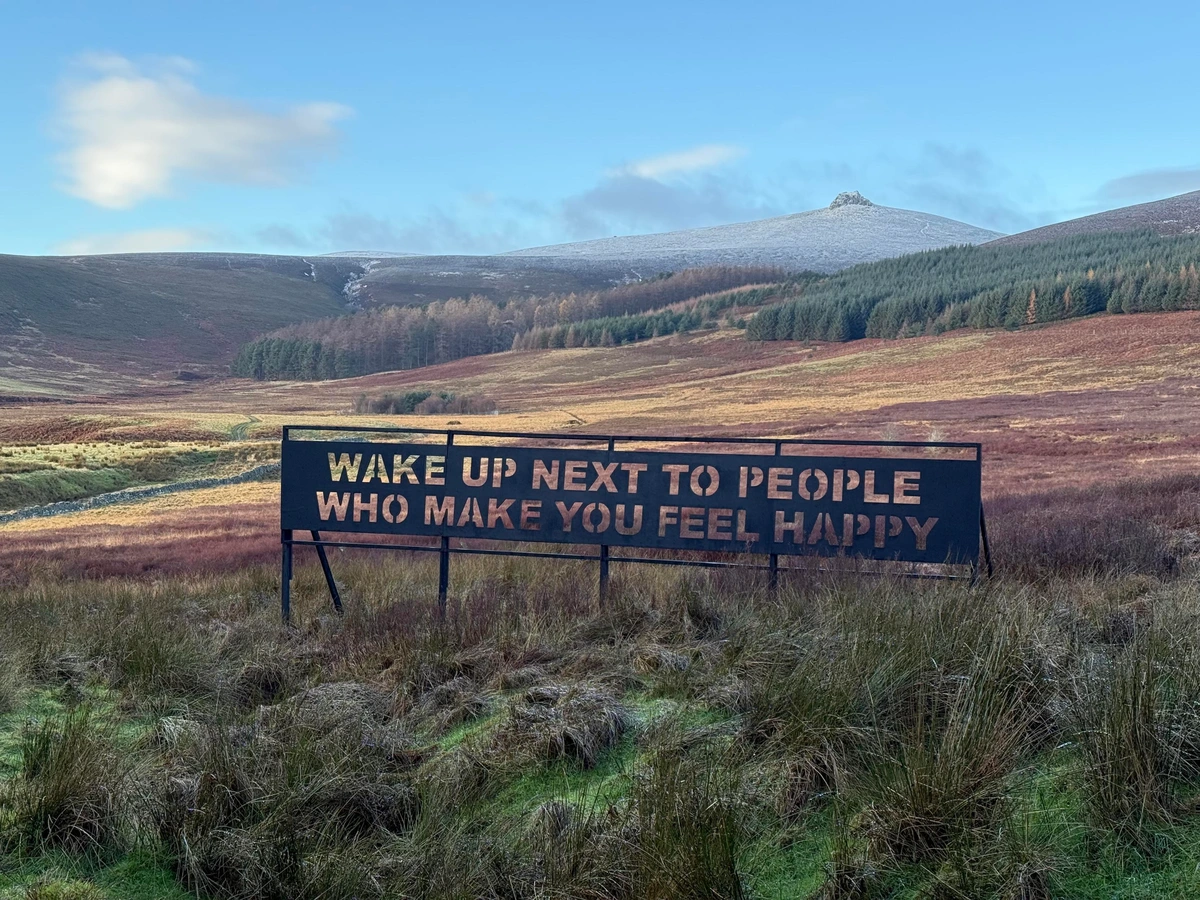 A large metal sign in a scenic landscape displays the text "WAKE UP NEXT TO PEOPLE WHO MAKE YOU FEEL HAPPY." The background features forests in the foreground, with the rocky peak of Clachnaben behind, partially covered in snow. 