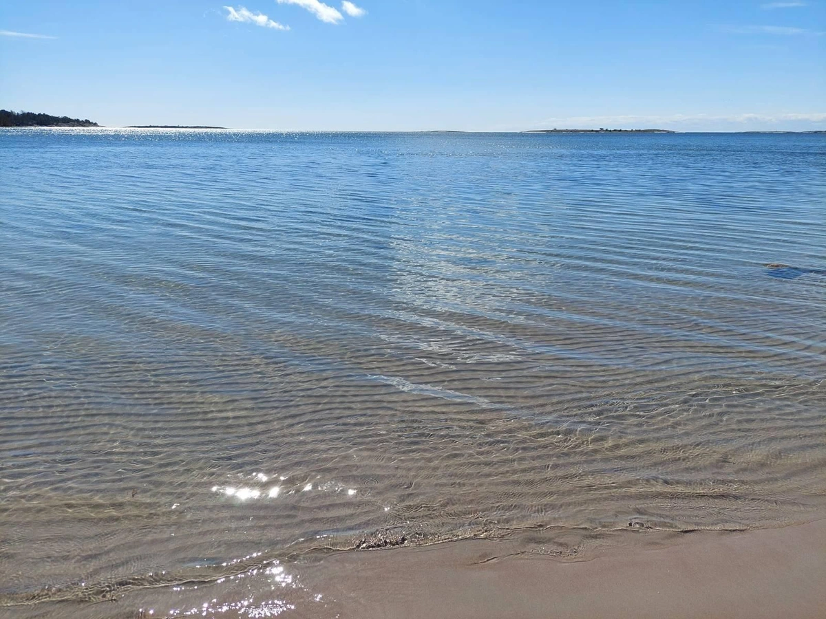 Photo of clear water showing waves in the sand made by the water, blue skies and a few islands in the background.