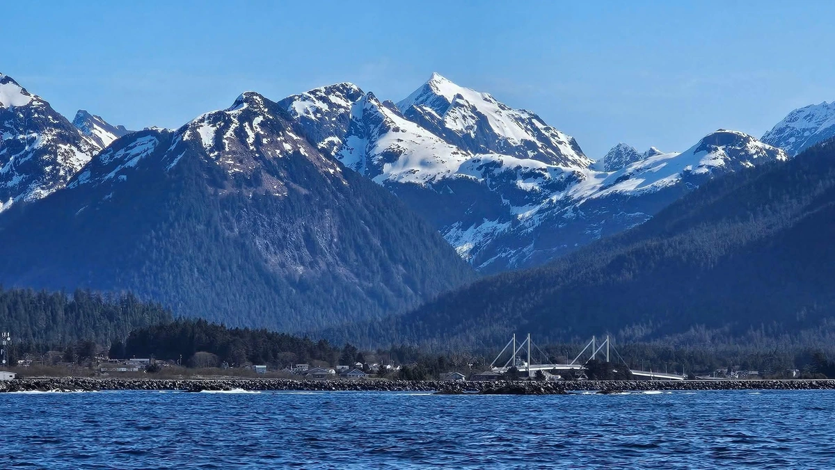 View of Sitka taken from a boat in Sitka Sound. The O'Connell, which connects Japonski and Baranof Islands in is the lower right, just above the water line. You can barely make out buildings along the shoreline, but huge, jagged mountains rise precipitously  in the shot, and the peaks are snow covered. The sky is cloudless and bright blue, giving the whole pic a blue cast when you add in the water. 