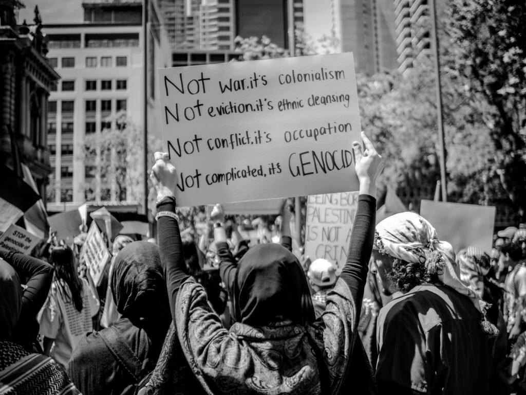 Protesters holding signs at a pro-Palestine rally, with one sign reading “Not war, it’s colonialism. Not eviction, it’s ethnic cleansing. Not conflict, it’s occupation. Not complicated, it’s genocide.”