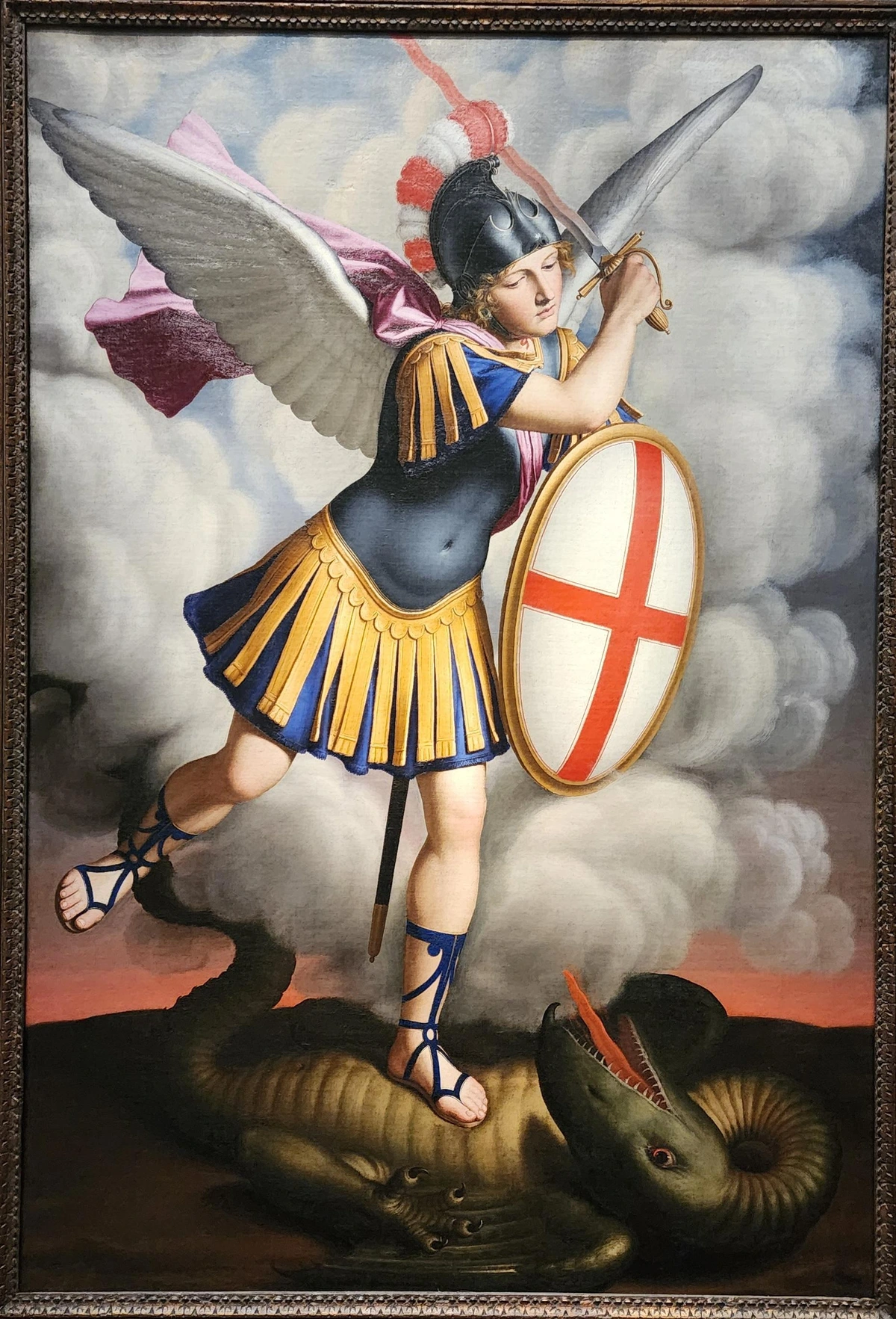 young caucasian male with wings wearing a bright blue and yellow skirted "Roman" style military uniform. he is holding a white shield with a red crossin front of him and a sword above his head, and he has a dragon under his left foot.