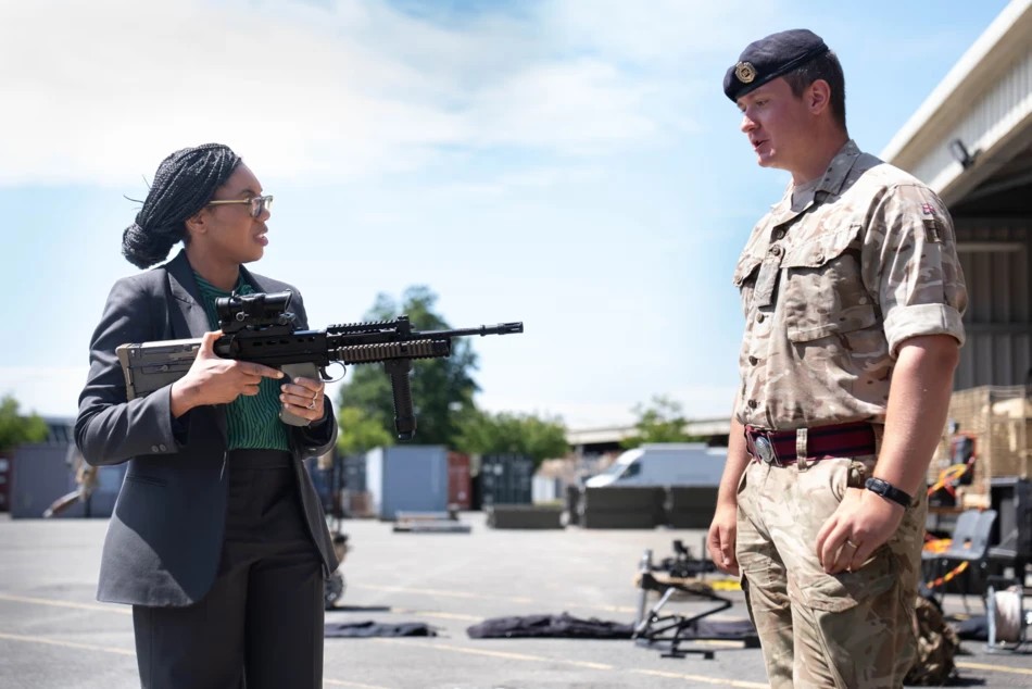 Kemi Badenoch holding an unloaded SA80 assault rifle pointed directly at a nervous looking solider with his thumps in his pockets.