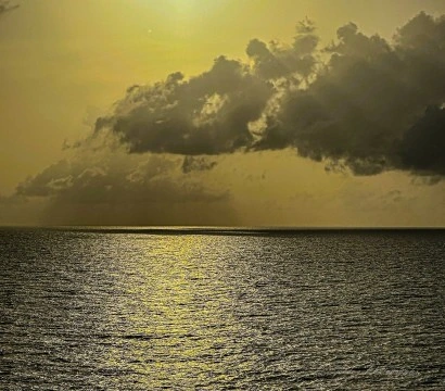 "The scene opens on the vast, open body of the Caribbean Sea—its surface a living tapestry of gentle ripples and shimmering light. The sun, low on the horizon, is either rising into promise or descending into memory. It casts a golden path across the water, like a celestial scroll unfurling toward the viewer. This radiant trail glows with molten amber, framed by the deep blues and silvers of the surrounding sea.

Above, the sky is a dramatic canvas of dark, brooding clouds—layered and textured like mythic veils. Some are tinged with firelight, their edges glowing as if kissed by the sun’s final or first breath. The contrast between light and shadow is striking: a sovereign interplay of warmth and mystery, clarity and concealment.

The air feels thick with reverence. There are no boats, no birds, no human interruptions—just the elemental majesty of water and sky in ceremonial dialogue. It’s a moment suspended in myth, where time pauses and the sea listens." - Microsoft Copilot