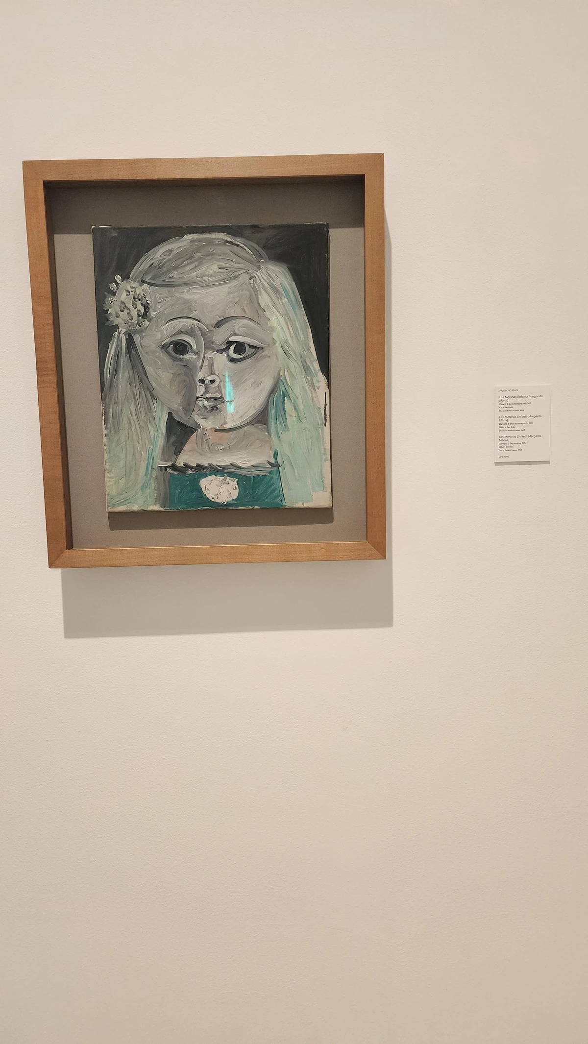A framed picture in light wooden frame hangs on museum wall. Picasso's Infanta Margarita María is part of his 1957 series of 58 variations on Velázquez's Las Meninas. Rendered in Cubist fragmentation, the young Spanish princess is reduced to bold geometric planes — angular white and grey forms suggesting her iconic wide skirt and pale face. Picasso strips away courtly realism, rebuilding her in flat, interlocking shapes that feel simultaneously playful and monumental. She is recognizable yet  reinvented — a dialogue across four centuries of painting.