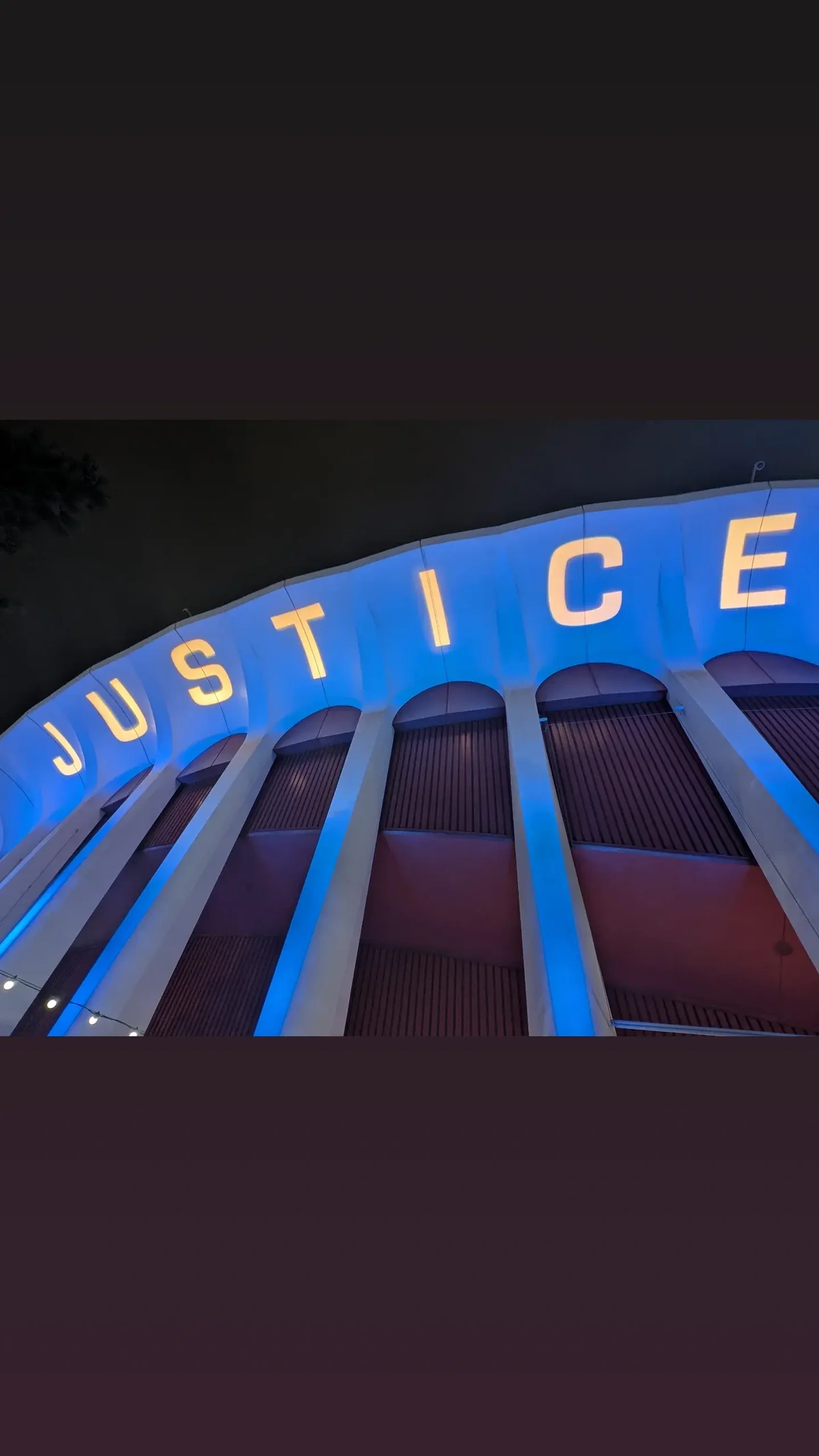 An image of the side of a stadium, with columns leading to a roof upon which JUSTICE is projected in white text with a blue background.
