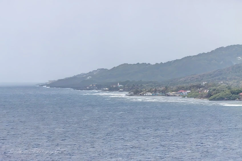 "From the vantage of open water, a Caribbean island rises gently into view, its shoreline curving like a welcoming arm. The sea in the foreground is calm and silvery, its surface textured with soft ripples that catch the muted light of an overcast sky. The horizon is low and wide, giving the scene a sense of spaciousness and quiet breath.

Along the coast, buildings nestle into the landscape—white walls, red roofs, and scattered structures that suggest homes, perhaps a church or lighthouse with a tower that punctuates the skyline. These human touches are modest and spaced, allowing the natural contours of the island to dominate.

Behind the shoreline, the land lifts into green hills, thick with trees and foliage. The vegetation is lush but not wild—more like a tended jungle, layered in shades of emerald and olive. The hills roll gently upward, their slopes softened by mist and distance.

The sky above is a soft gray, blanketing the scene in a quiet hush. There’s no drama here—just a serene meeting of sea, land, and sky. The image captures a moment of stillness, as if the island is pausing between tides, waiting for the next breeze or boat to arrive." - Microsoft Copilot