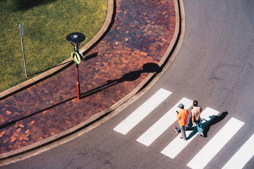The image shows an aerial view of two people walking across a zebra crossing on a road. Next to the road is a brick-paved sidewalk and a grassy area. There is a pole with signs casting a shadow on the ground.