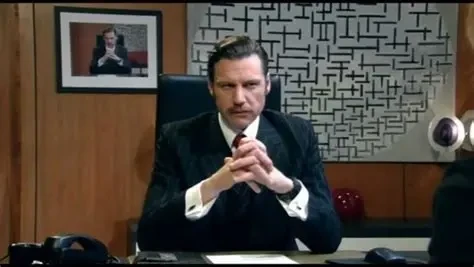 The IT Crowd - Denholm Reynholm sat at desk in front of a portrait of him sat at desk