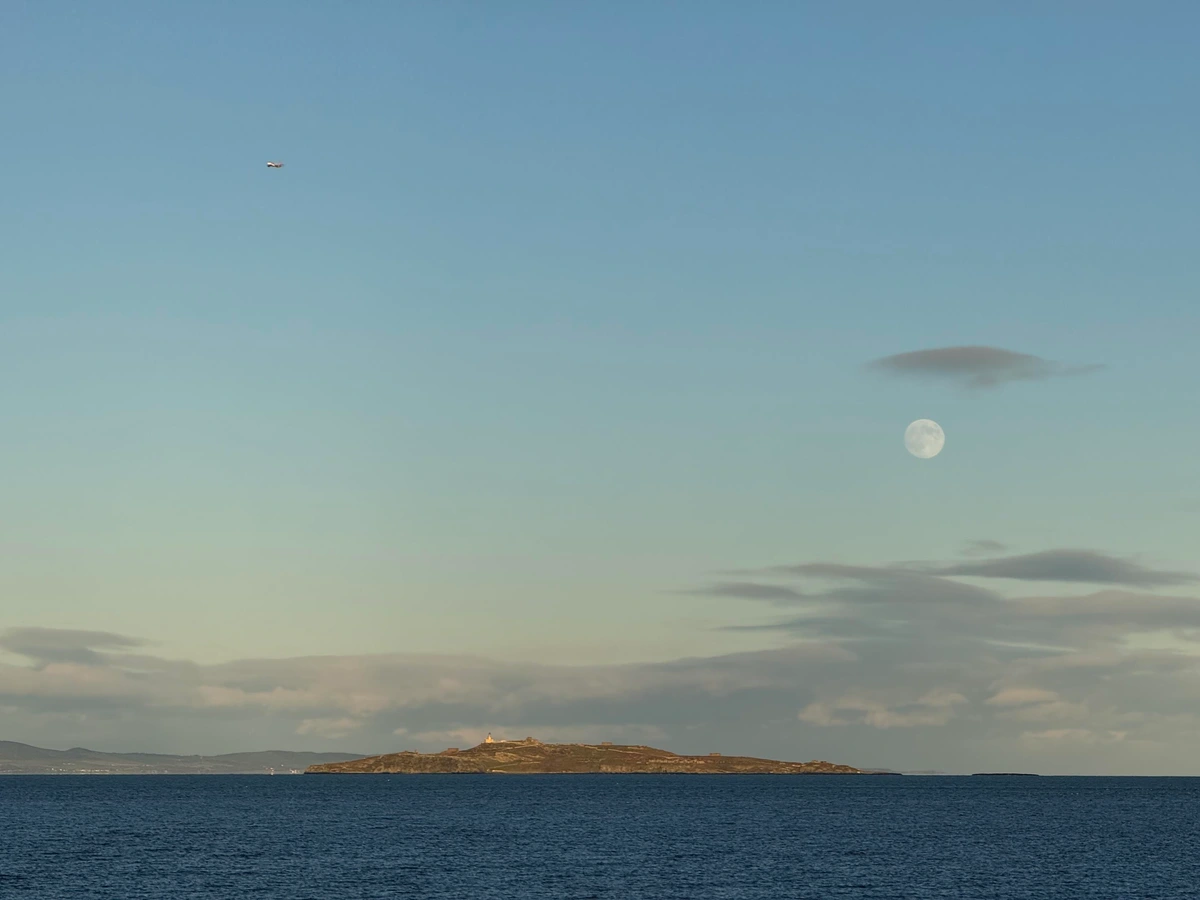 The island of Inchkeith in the calm deep blue of the Forth. Above, a blue sky with a few scattered clouds near the horizon. The almost-full moon is visible to the right of the island. 