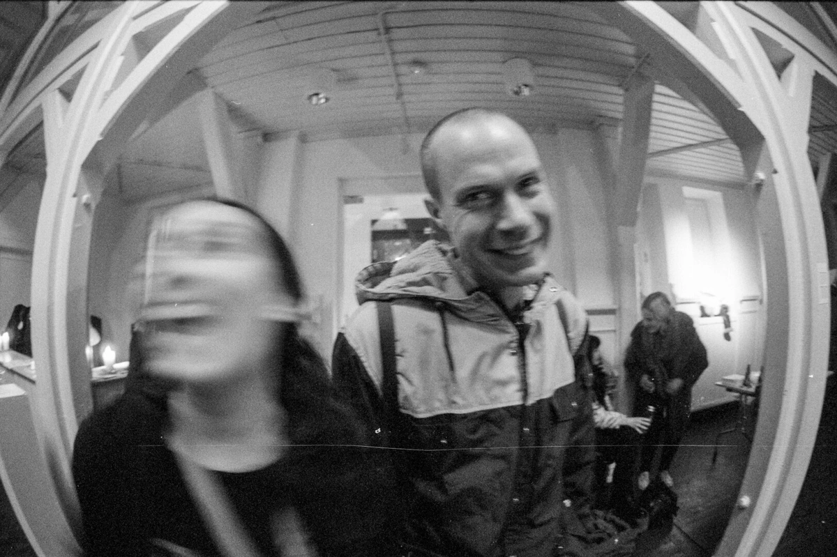 Closeup photo of two persons, the one two the left is moving her head and is a bit fuzzy on the photo, the man on the right is smiling happily. Some people are drinking in the background. Looks like a gallery space.