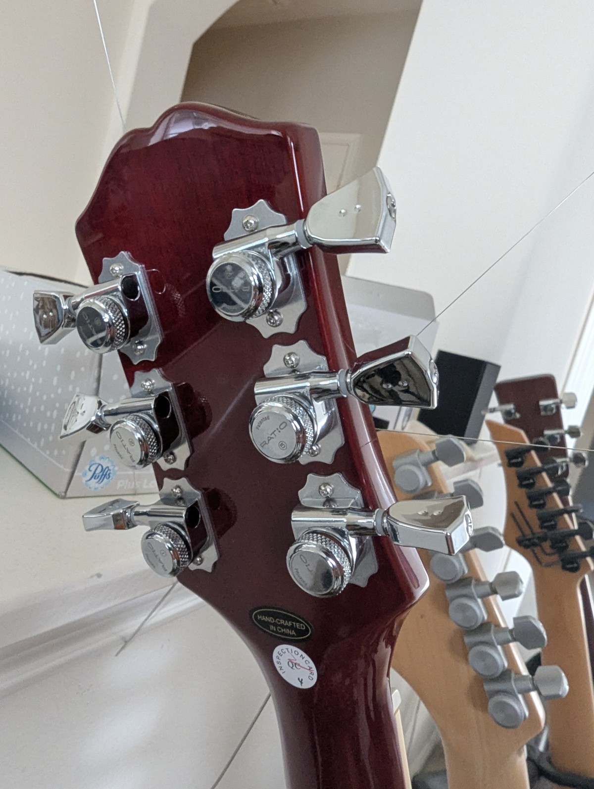 The foreground subject is the back of the headstock on an Epiphone ES-335 guitar. it features shiny chrome locking tuners by Graphtech. In the background are the headstocks from three other guitars: a Fender Stratocaster; an Ibanez style, and an acoustic.