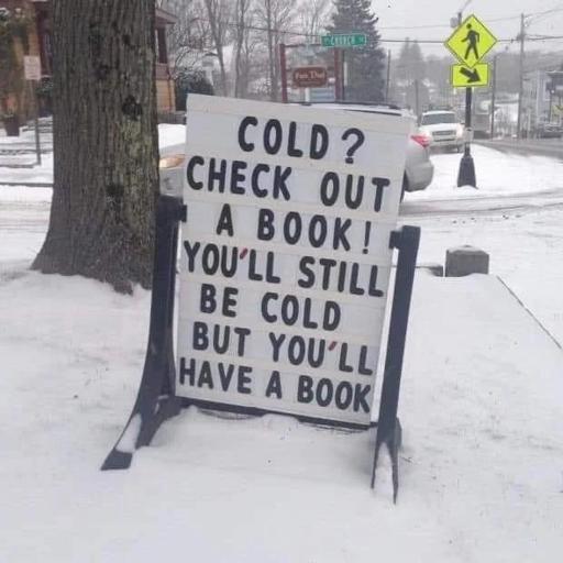 A white sign with black lettering standing a bit crooked in the snow saying, Cold? Check out a book! You'll still be cold but you'll have a book!