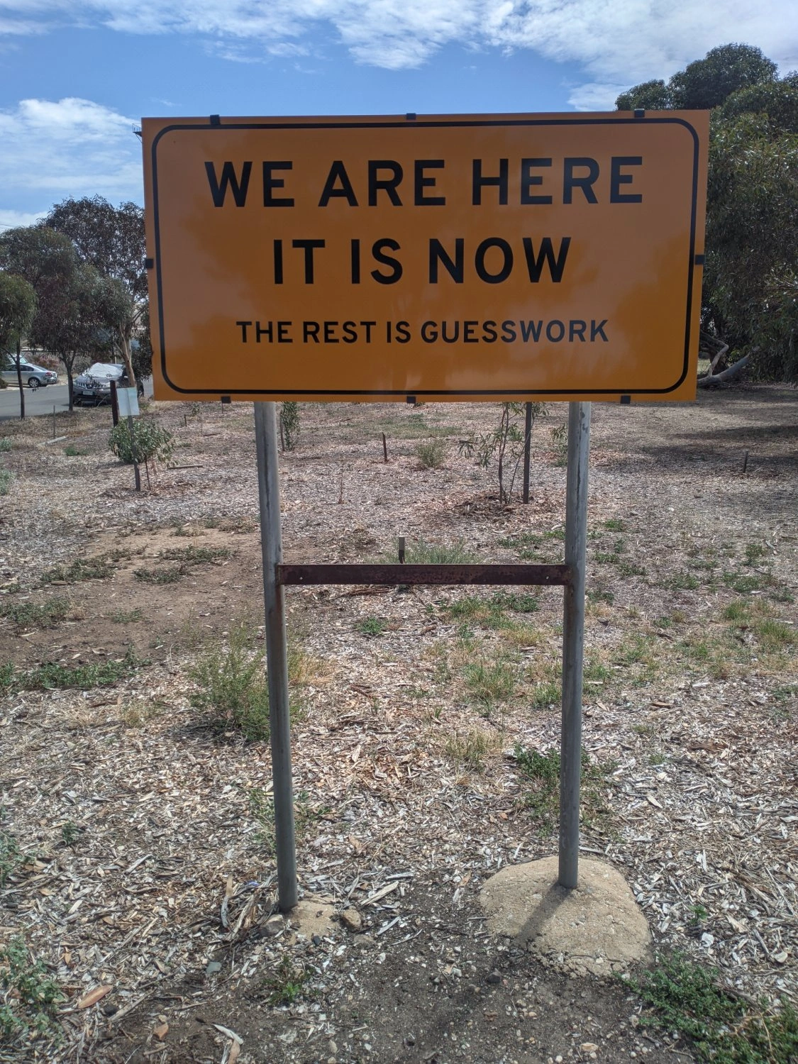A yellow roadside-like sign that says 'We Are Here. It Is Now. The Rest Is Guesswork'