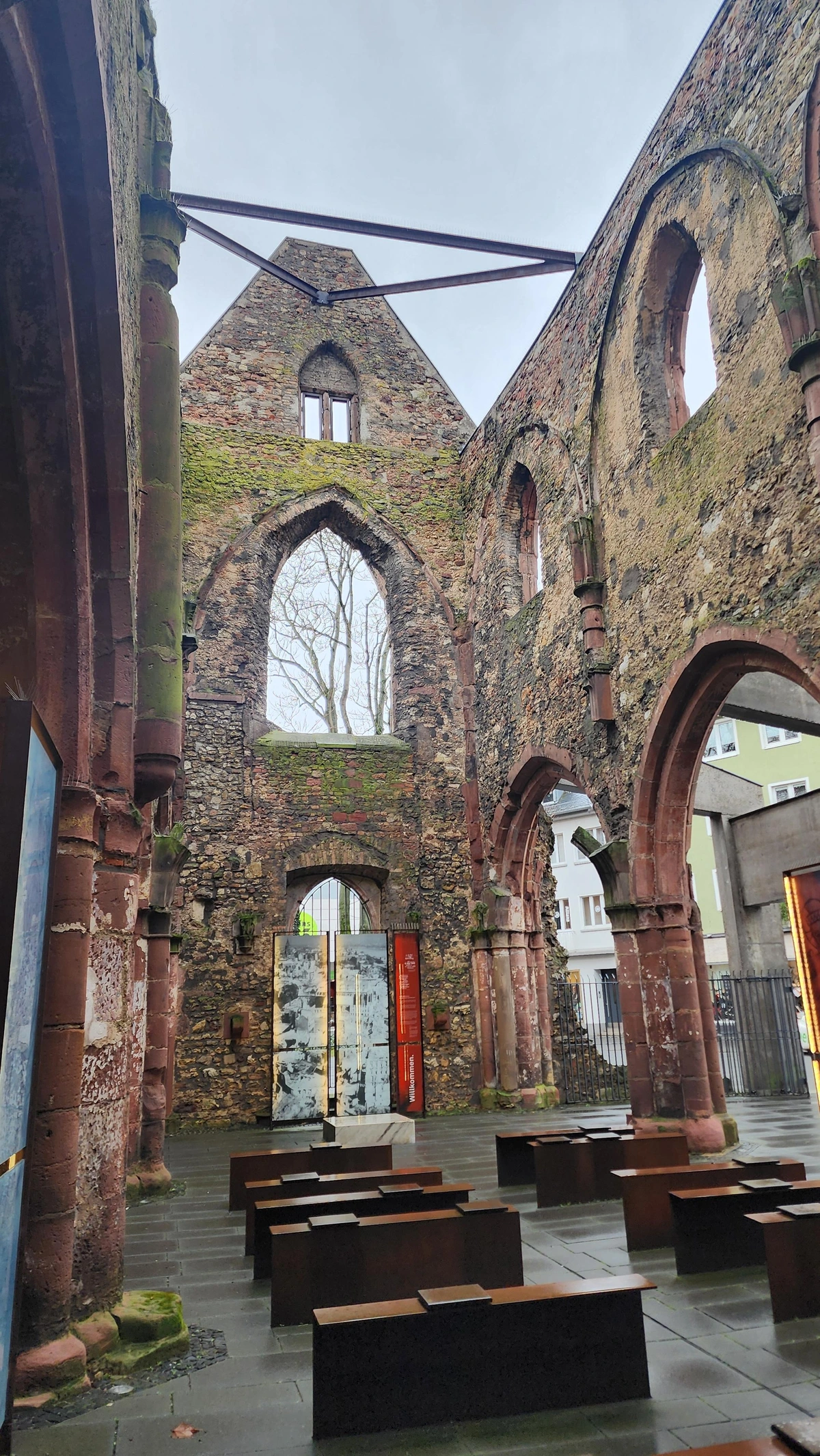 Inside the skeletal ruins of a smallish red stone church, looking from the altar to the entrance. Only the arches and upper windows of the nave and the back wall remain. The floor is concrete with modern, minimalist "pews" made of old steel beams. modern buildings are visible between the arches, and the overcast sky is visible through the roofless ceiling.
