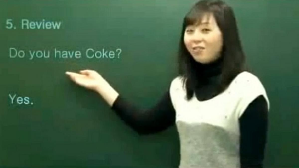korean language teacher in front of chalk board with question written about asking do you want coke? and looking at viewer png