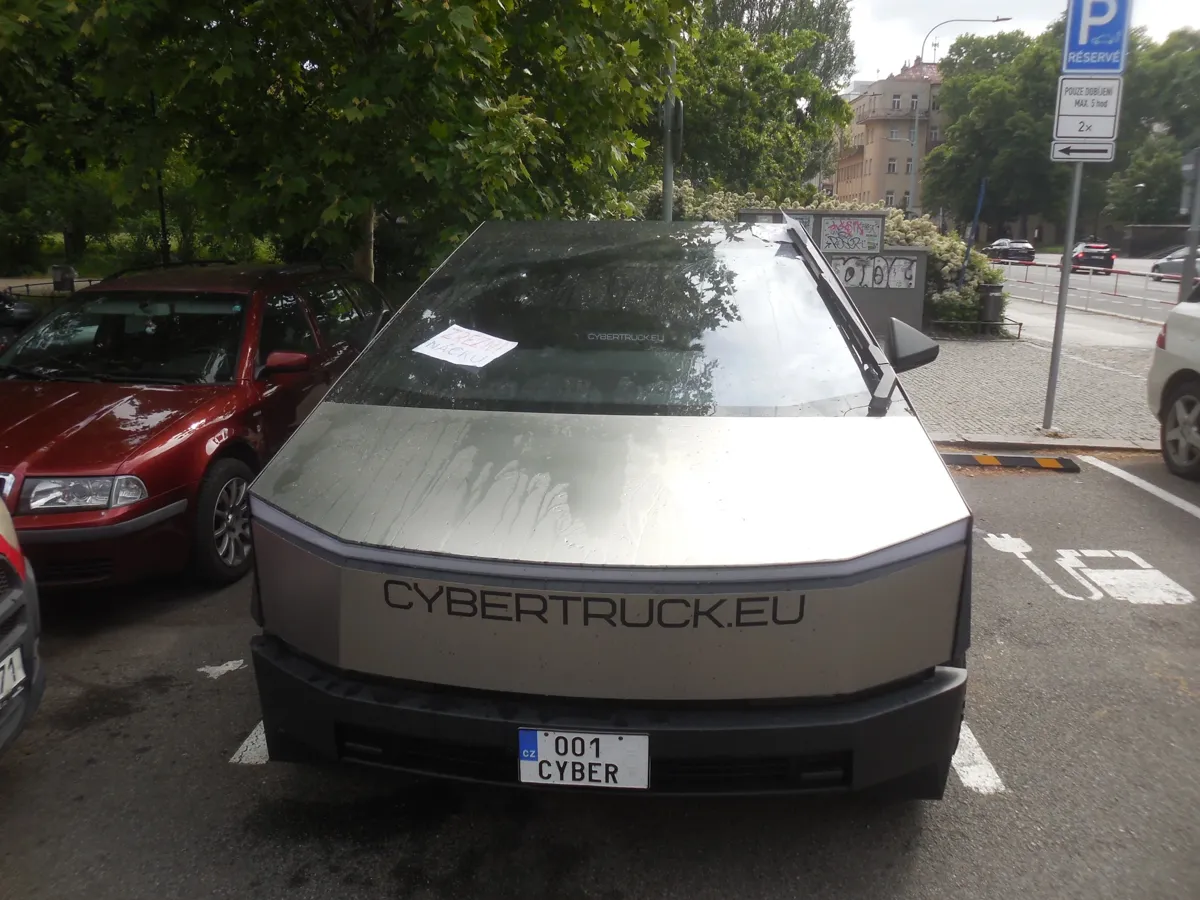 Tesla Cybertruck with custom near-invisibly rounded edges to pass EU standards, charging at a parking lot in central Prague. It is monstrous compared to a 2000s family car next to it. Giant "CYBERTRUCK.EU" sticker on the front. Licence plate: "001 CYBER". The weather is sunny but the car has been splashed with water, with a sheet of paper left on the windshield.