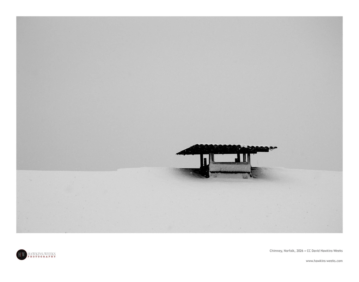 Black and white photo of a chimney and cowl on the right, rising above a snow-covered roof against a grey, cloudy sky
