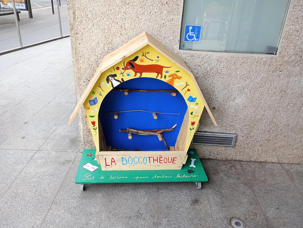 Photo of some kind of handmade "bookcase" for storing wooden sticks, painted with cute dog pictures and bearing these writings in French: "la doggothèque", "Prêt de bâtons pour toutous lecteurs".