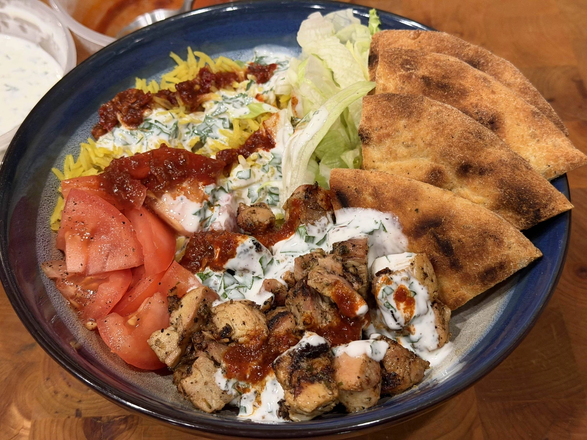 A bowl of yellow turmeric rice with shredded lettuce, sliced tomatoes, naan bread wedges, and grilled chicken pieces. Topped with a mayo and vinegar white sauce and harissa red sauce.