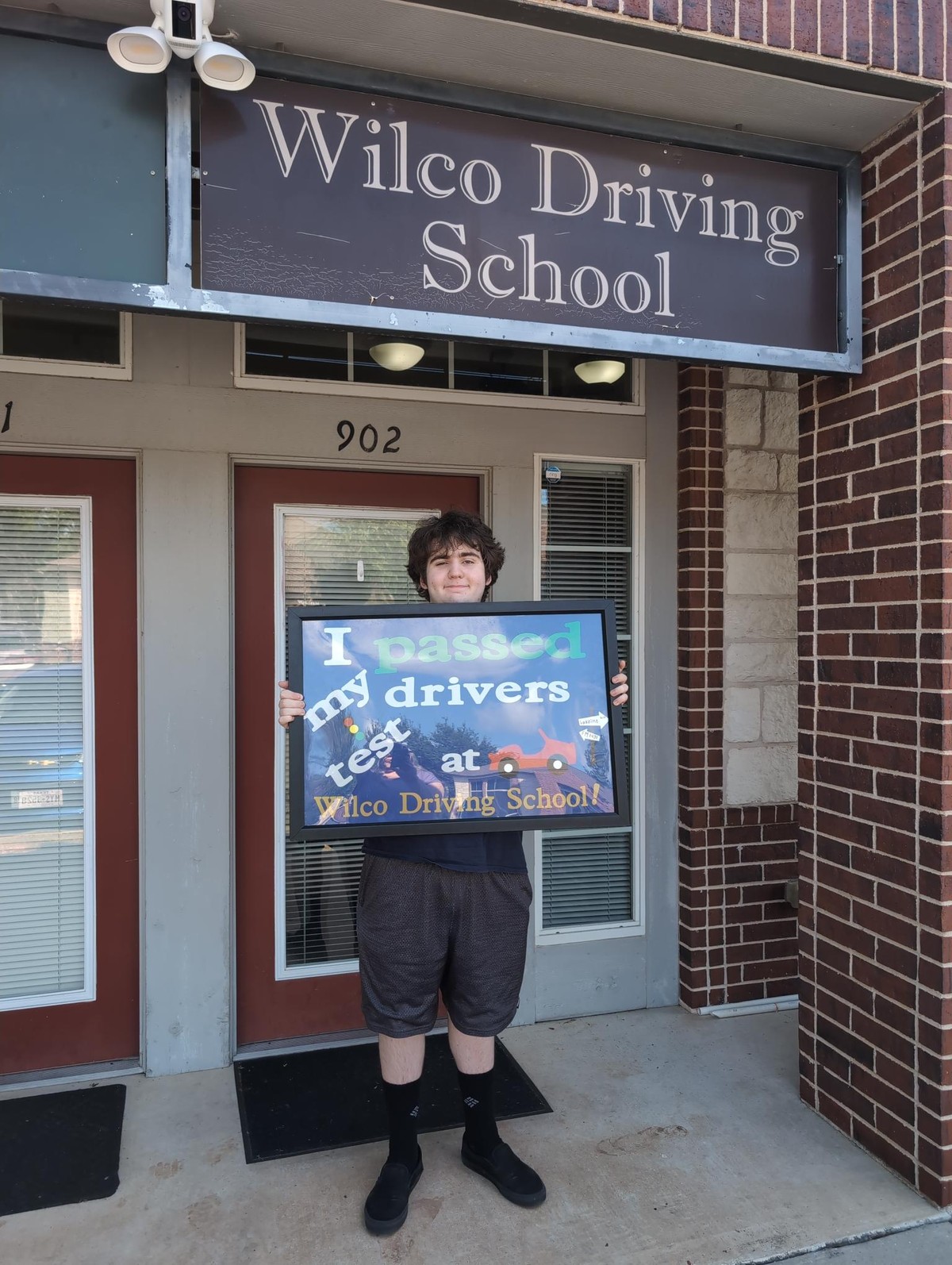 My 18 year old standing holding a sign that says "I passed my drivers test at Wilco Driving School!"