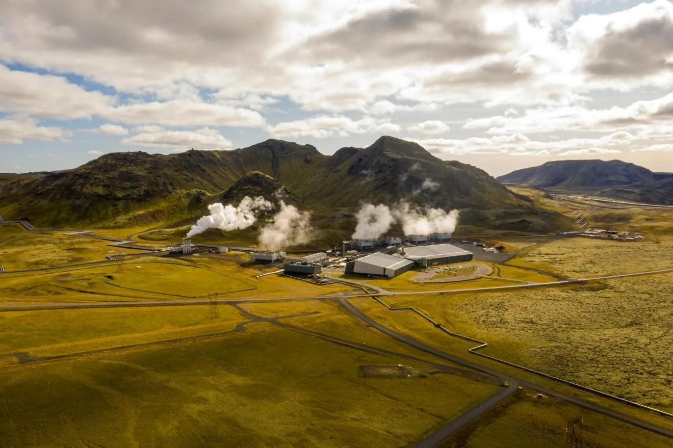 Photo from the air of a landscape showing fields with short mountains. In front of the mountains is a geothermal plant.