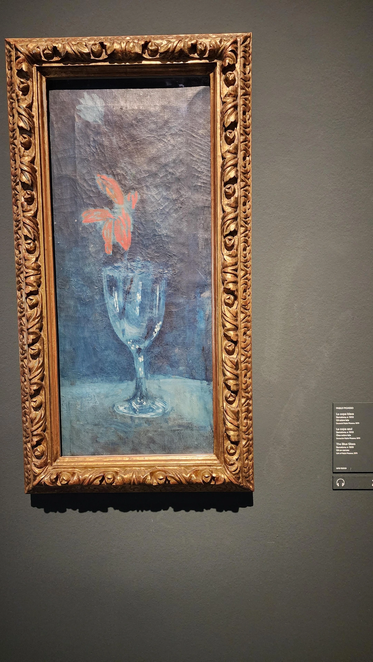 A tall, narrow canvas — roughly twice as tall as wide — painted around 1903 during Picasso's Blue Period. At the center stands a single transparent drinking glass holding one red flower. The petals are the only warm note; everything else — the glass, the background, the surrounding space — is suffused in deep, cool blue. The composition is spare and still, almost meditative. Blue doesn't just surround the flower; it seems to absorb it.