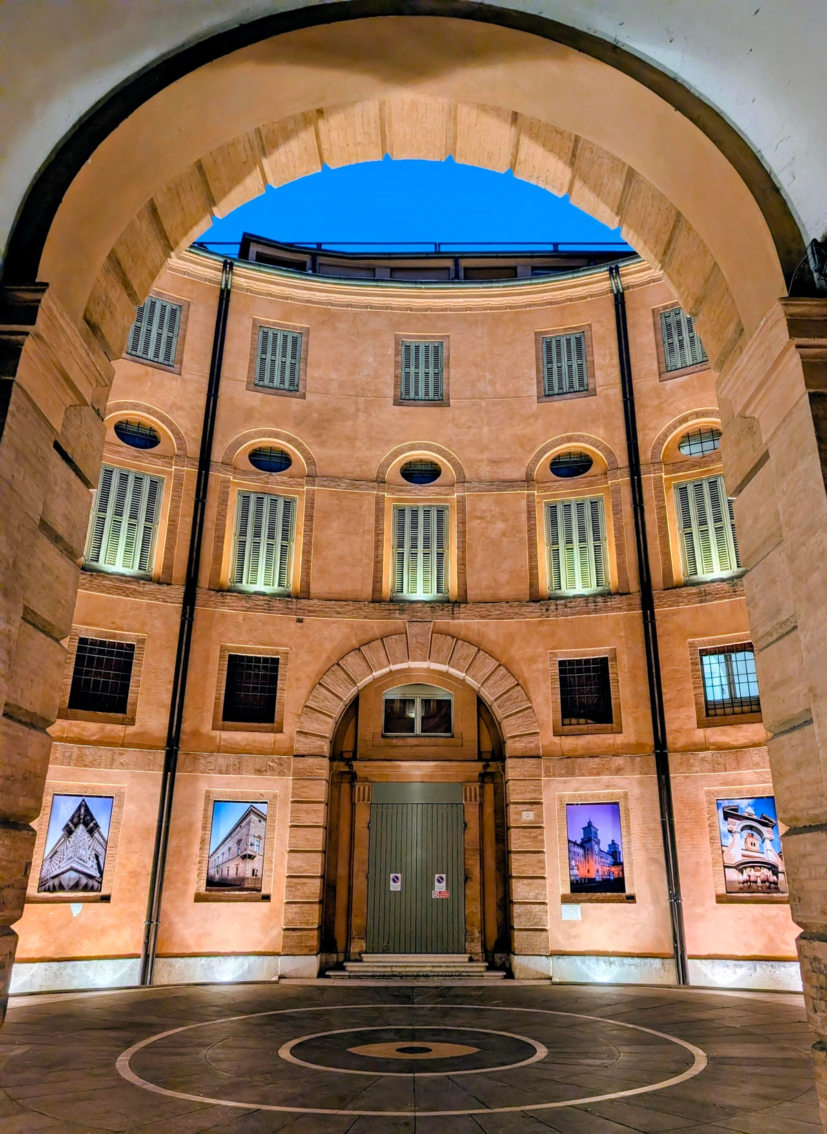A low-angle shot captures the symmetrical, curved facade of an illuminated historic building at night, viewed through a large archway. The building's terracotta walls are lit from below, highlighting multiple windows with shutters and four framed artworks. The circular patterned paving in the foreground emphasizes the curved architecture, all beneath a deep blue night sky visible through the arch.