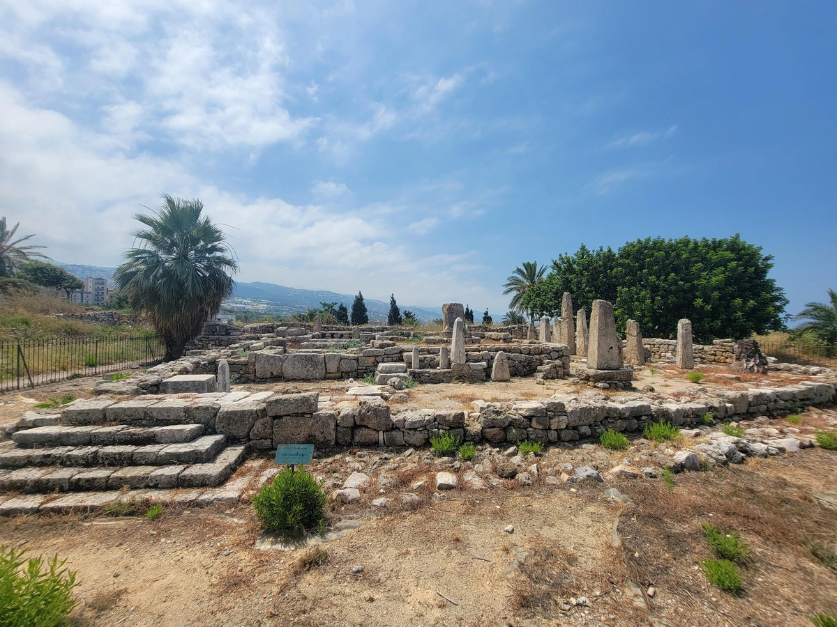 A seaside hill with shrubs and small trees under a slightly cloudy sky. At center a large stone square platform with 2 or 3 successively smaller, higher levels. Interspersed on the different levels are stone obelisks of various sizes and states of erosion.  At left is a restored or well-preserved set of 4 steps leading up to the lowest platform.
