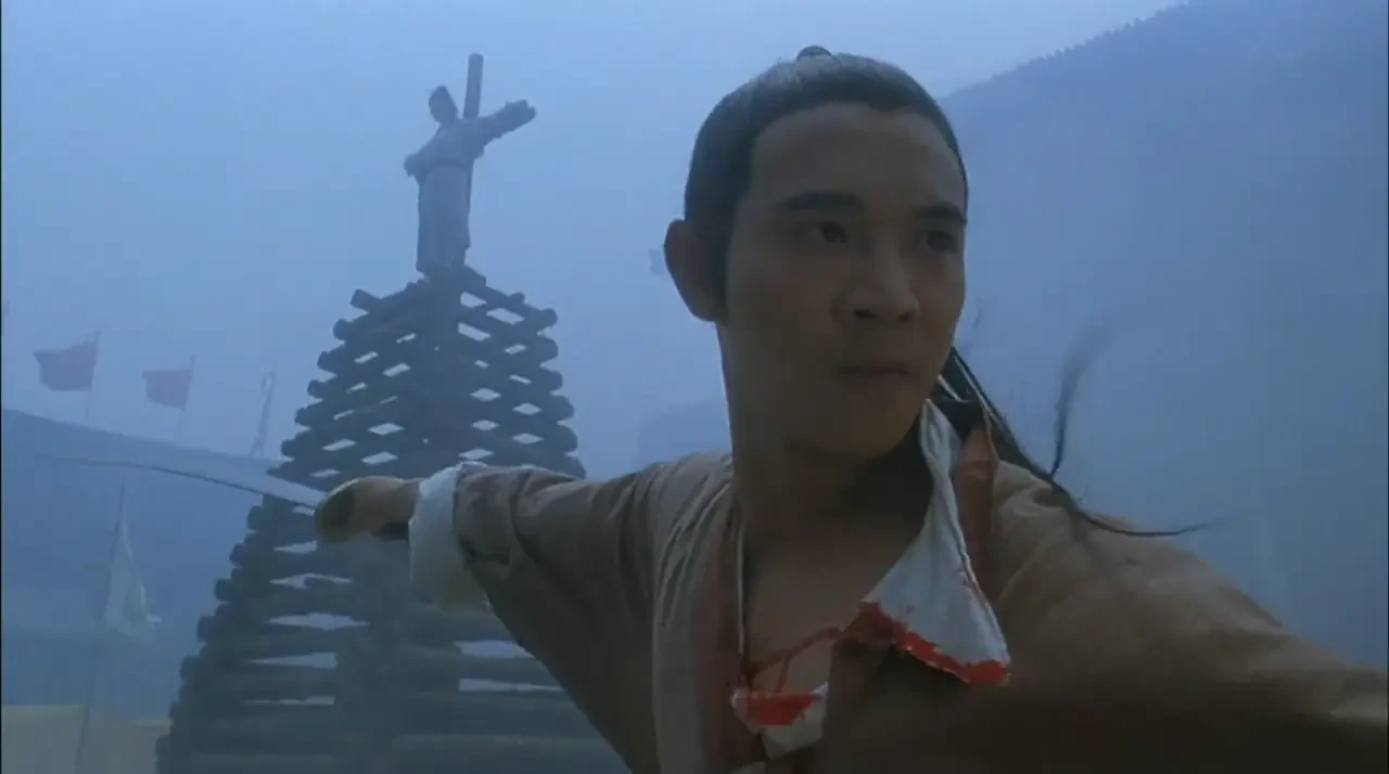 A man holding a sword is in a fighting stance. Behind him, a structure of logs with atop a person tied to a cross.