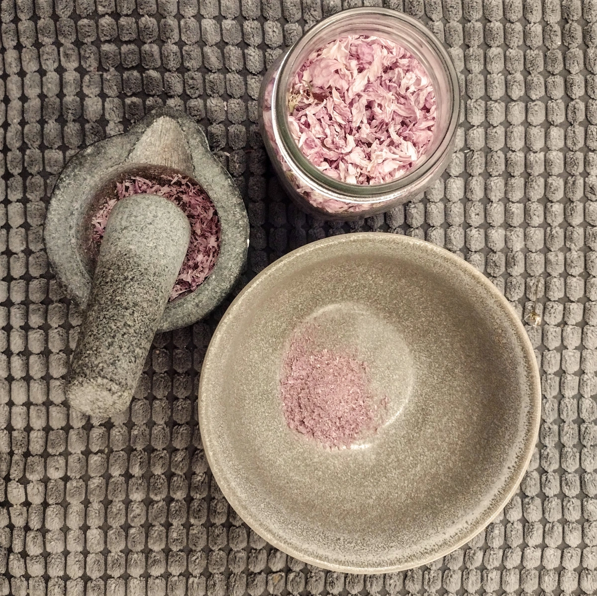 A top-down view of a glass jar of dried cherry blossoms, a mortar and pestle, and pulverized blossom power in a bowl, on a gray fabric background.