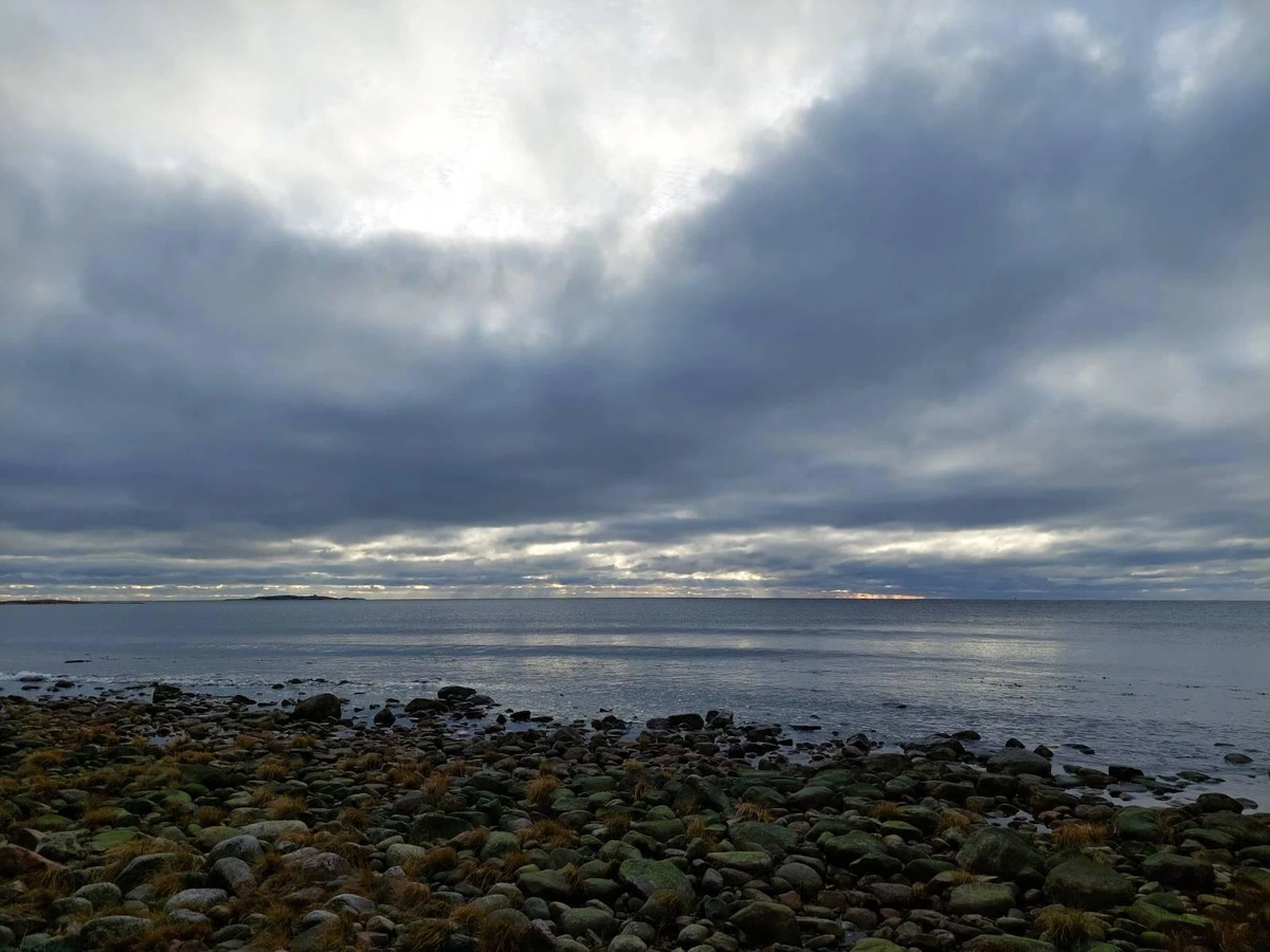 Photo of a stoney shore with grey clouds over the water, a couple of islands in the distance, and a little bit if light making its way through the clouds.