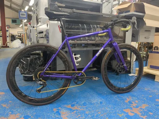 Purple Ragley Trig bike with gold chain and carbon wheels sitting in front of industrial machinery. 