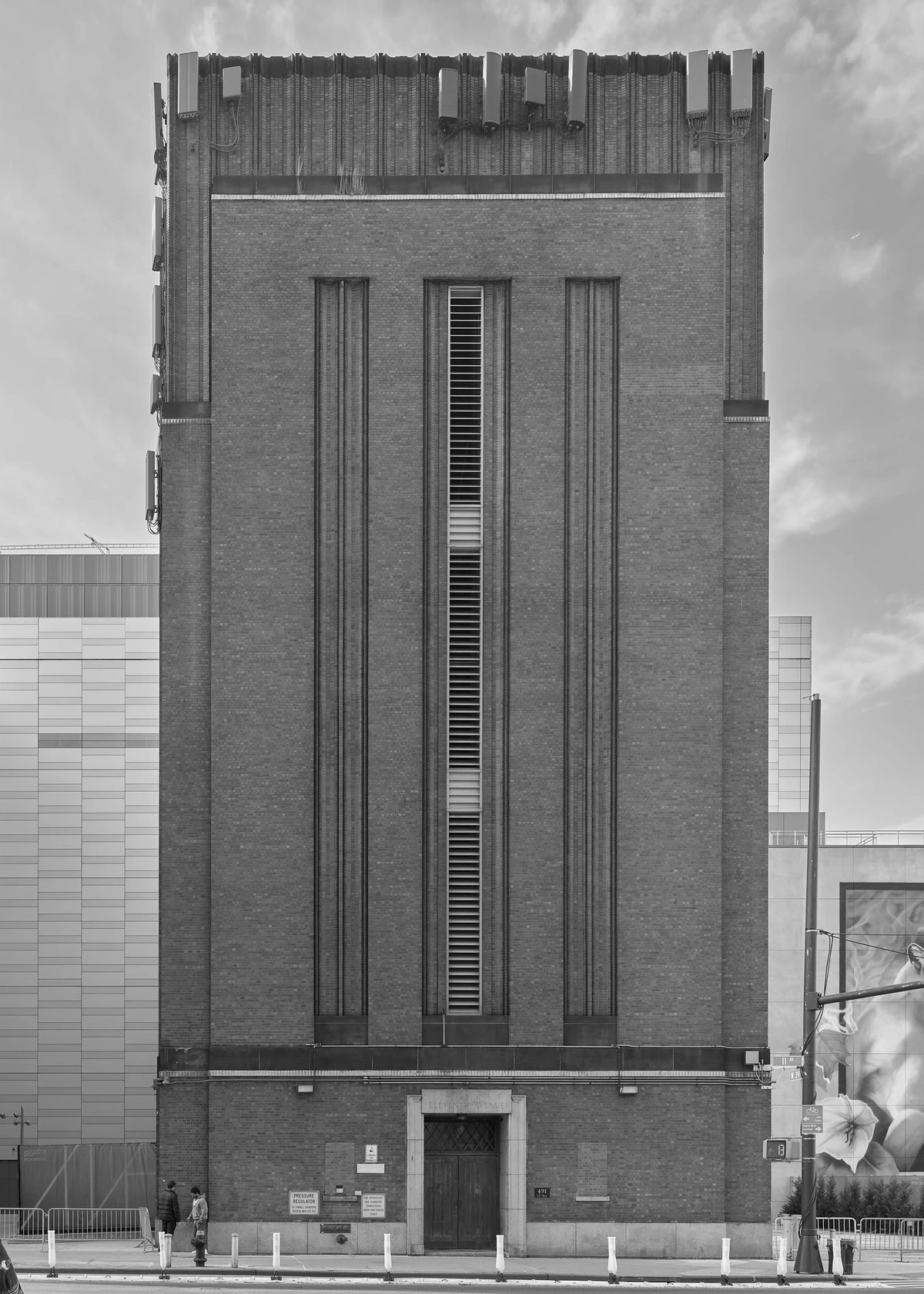 A simple industrial art deco-style building, lacking windows, but with a narrow louvered vent at center running vertically along its length, on a city street.