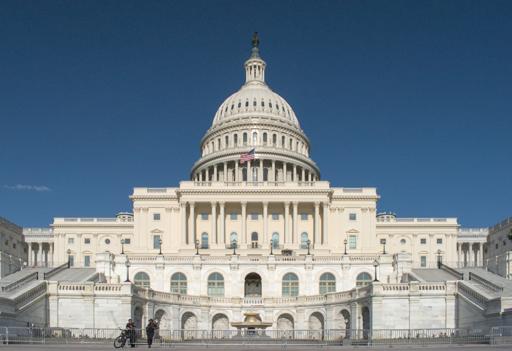 A photo of the US Capitol, from the west side, during a sunny day with clear skies, a couple of capitol cops stand by on metal barricades in front, one has a bike. The sun shines on this side of the building.
